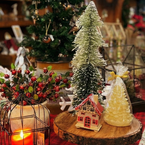 Festive holiday display featuring a red candle in a metal lantern, berry stems, miniature Christmas trees, and a small decorative house arranged on a red plaid tablecloth.