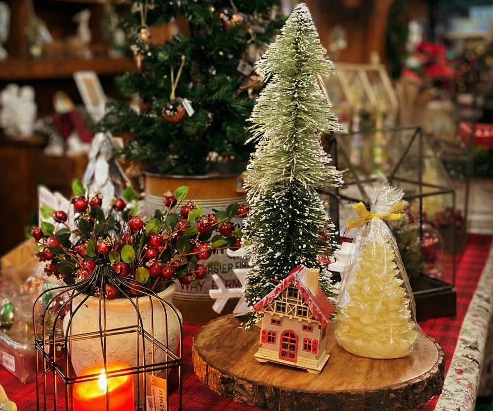 Festive holiday display featuring a red candle in a metal lantern, berry stems, miniature Christmas trees, and a small decorative house arranged on a red plaid tablecloth.